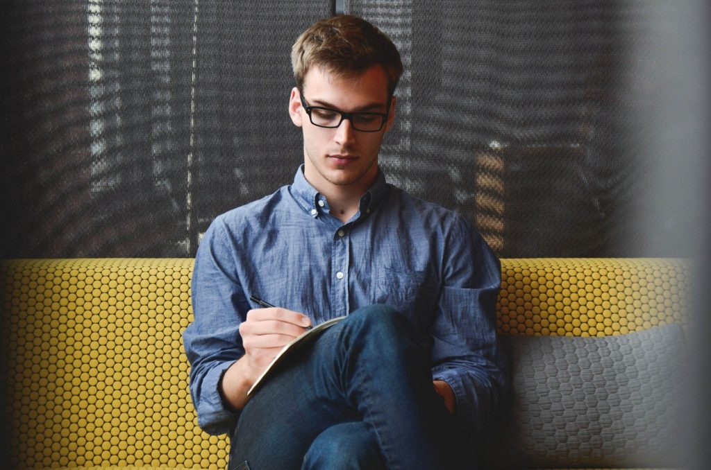 un jeune homme à lunettes sur un canapé tient un stylo et un carnet sur lequel il semble prendre des notes.